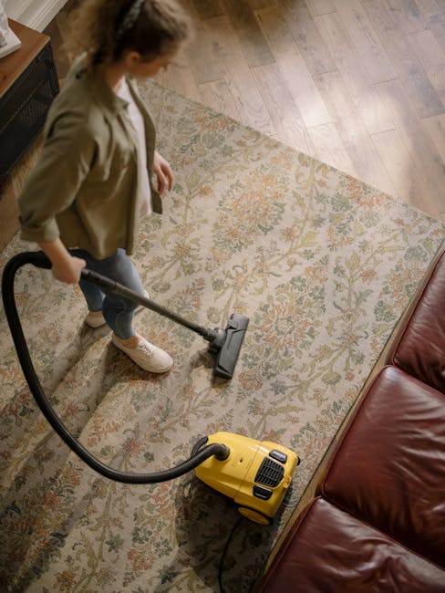 A woman is performing surface cleaning on a traditional patterned area rug with a yellow vacuum cleaner in a living room. The rug features intricate floral designs in muted tones and is placed on a wo