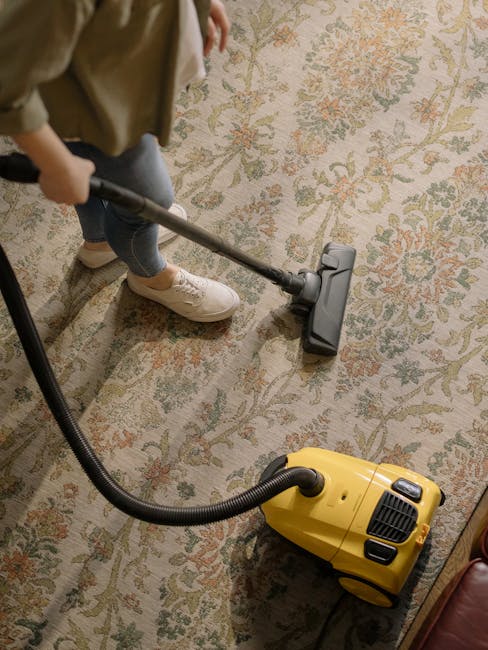 A woman is performing surface cleaning on a traditional patterned area rug with a yellow vacuum cleaner in a living room. The rug features intricate floral designs in muted tones and is placed on a wo