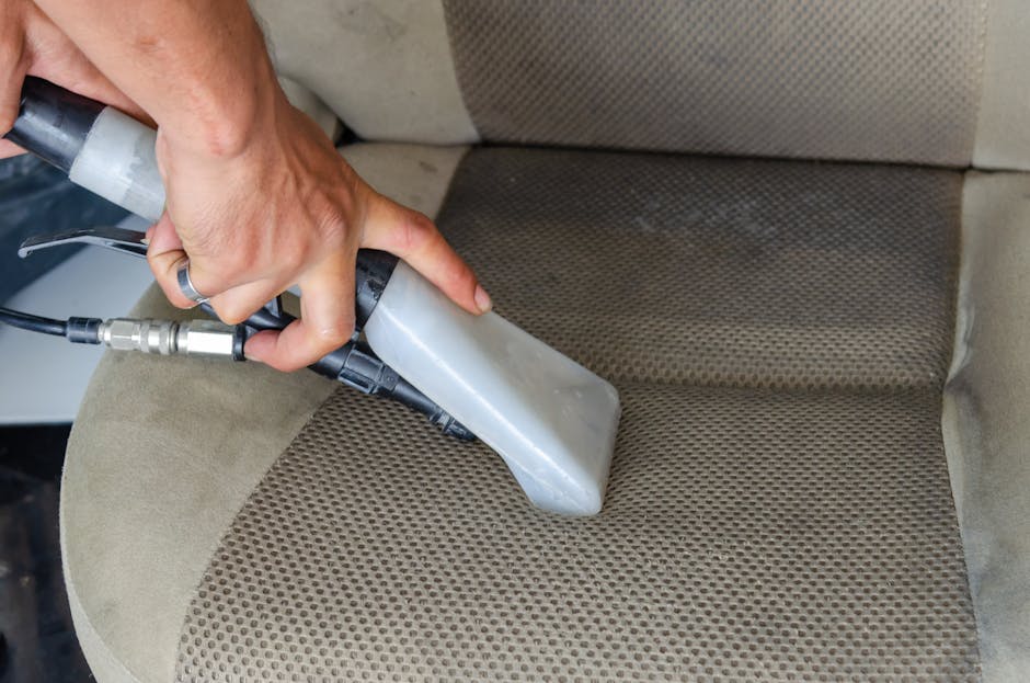 A person dressed in full protective cleaning gear, including a white suit, gloves, and a face mask, is using a vacuum or fogging device to sanitize a living room. The room has a beige sofa with patter