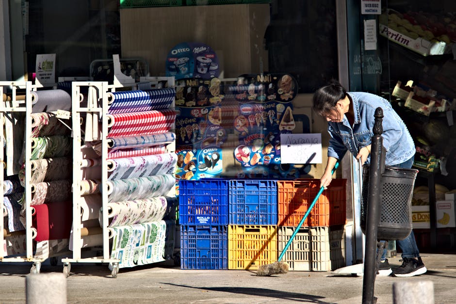 A woman wearing a blue denim jacket and black trousers is engaged in surface cleaning outside a shop at Woolwich Market, as part of a case study by Woolwich Carpet Keepers. She is using a broom to swe