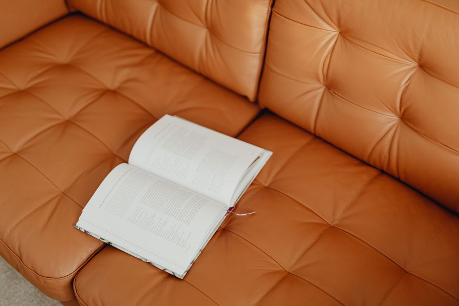 A close-up view of a tan leather sofa in a well-lit living room, showing a smooth, shiny surface with visible tufted buttons on the backrest and seat cushions. An open hardcover book with white pages
